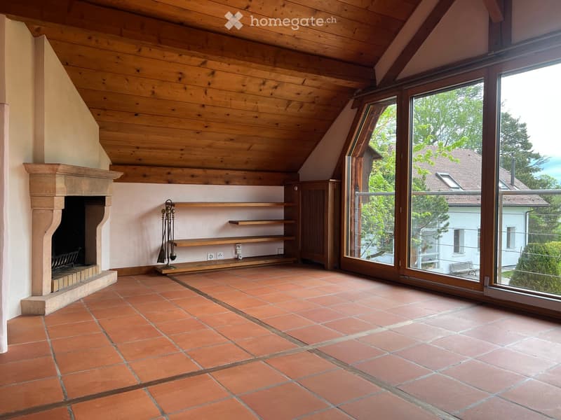 An unfurnished room with exposed wooden beams, terracotta tiles, a fireplace, and large windows offering a view of the outdoors.