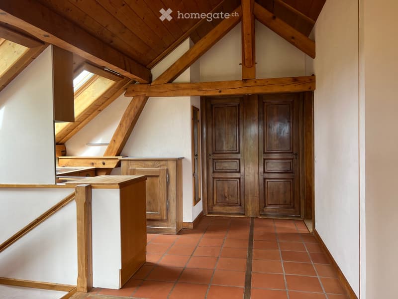 A spacious, well-lit hallway with exposed wooden beams and a high, slanted ceiling. The floor is covered in terracotta tiles, and the walls are painted white. There are two wooden double doors at the end of the hallway, and a wooden bench or table is posi
