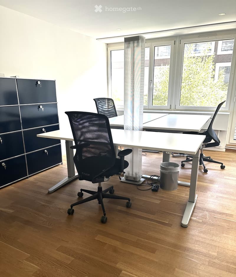 Office space with wooden flooring, large windows, white table, black office chairs, and a dark cabinet.