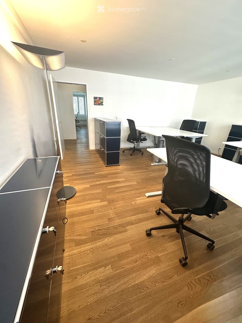 Modern office space with wooden flooring, white tables, black office chairs, and a sleek cabinet.
