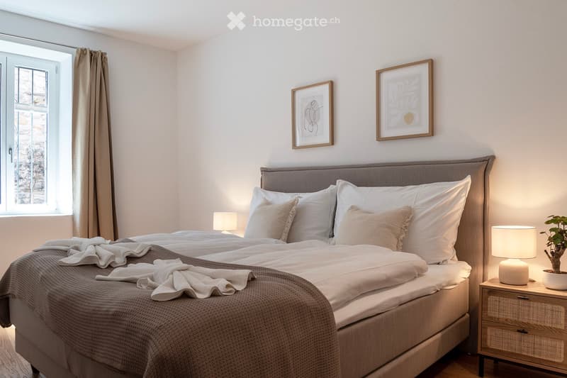 Spacious bedroom with large window, gray upholstered bed frame, white bedding, gray throw blanket, two framed artwork on the wall, and a bedside table with lamps