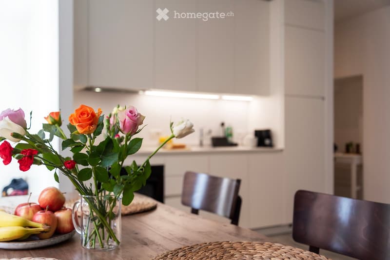 Wooden dining table with a vase of colorful roses, a bowl of fresh fruits, and a modern kitchen with shelves and appliances in the background