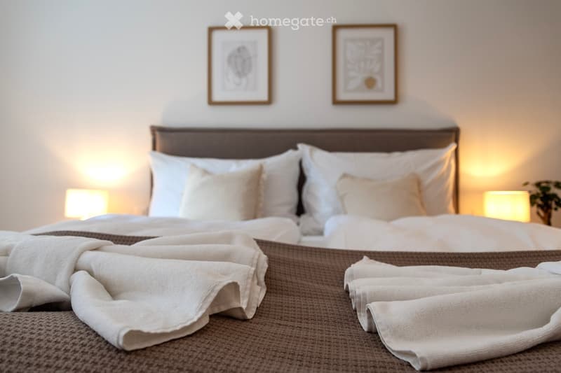 Cozy bedroom with a large bed, white bedding, and a brown woven blanket. Two framed artwork pieces hang on the wall above the bed. The room has a warm, inviting atmosphere with soft lighting from the lamps on the nightstands.