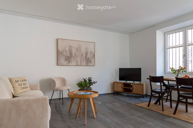 Spacious living room with gray walls, hardwood floors, large windows, a sofa, a coffee table, a TV stand, and a dining table with chairs. The room also features a piece of abstract art on the wall and a potted plant.