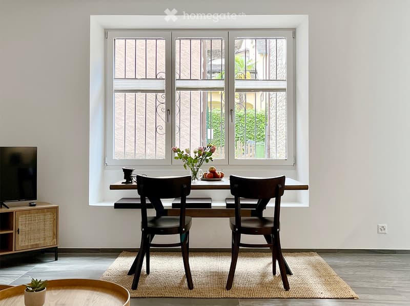 Dining room with large windows, wooden dining table and chairs, TV stand, and a vase of flowers on the table.