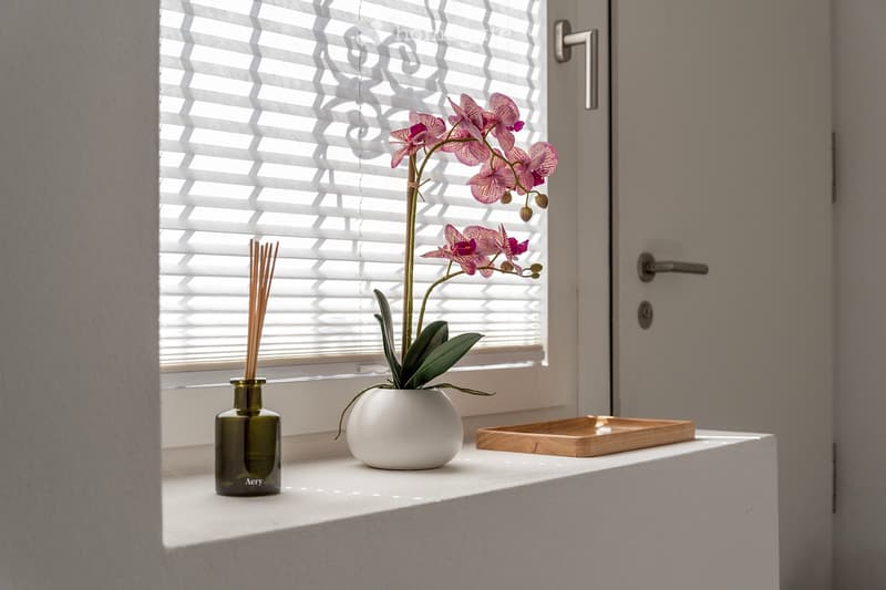 The image shows a room with a window, a white vase containing a pink orchid plant, a glass bottle with a diffuser, and a wooden tray on a shelf.
