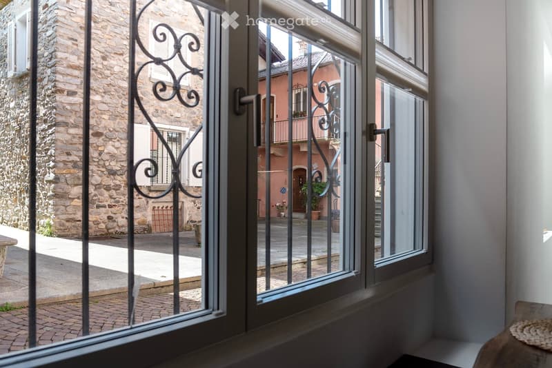 The image shows the interior of a building with large windows and a wrought iron gate. The walls are made of exposed stone, and there is a balcony or terrace visible through the windows.