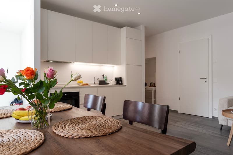 Modern kitchen with white cabinets, wooden dining table, woven placemats, and a vase of colorful flowers