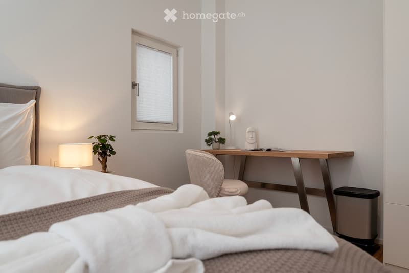Cozy bedroom with a large window, wooden desk, and decorative plants. The room has a neutral color scheme and appears to be well-lit.