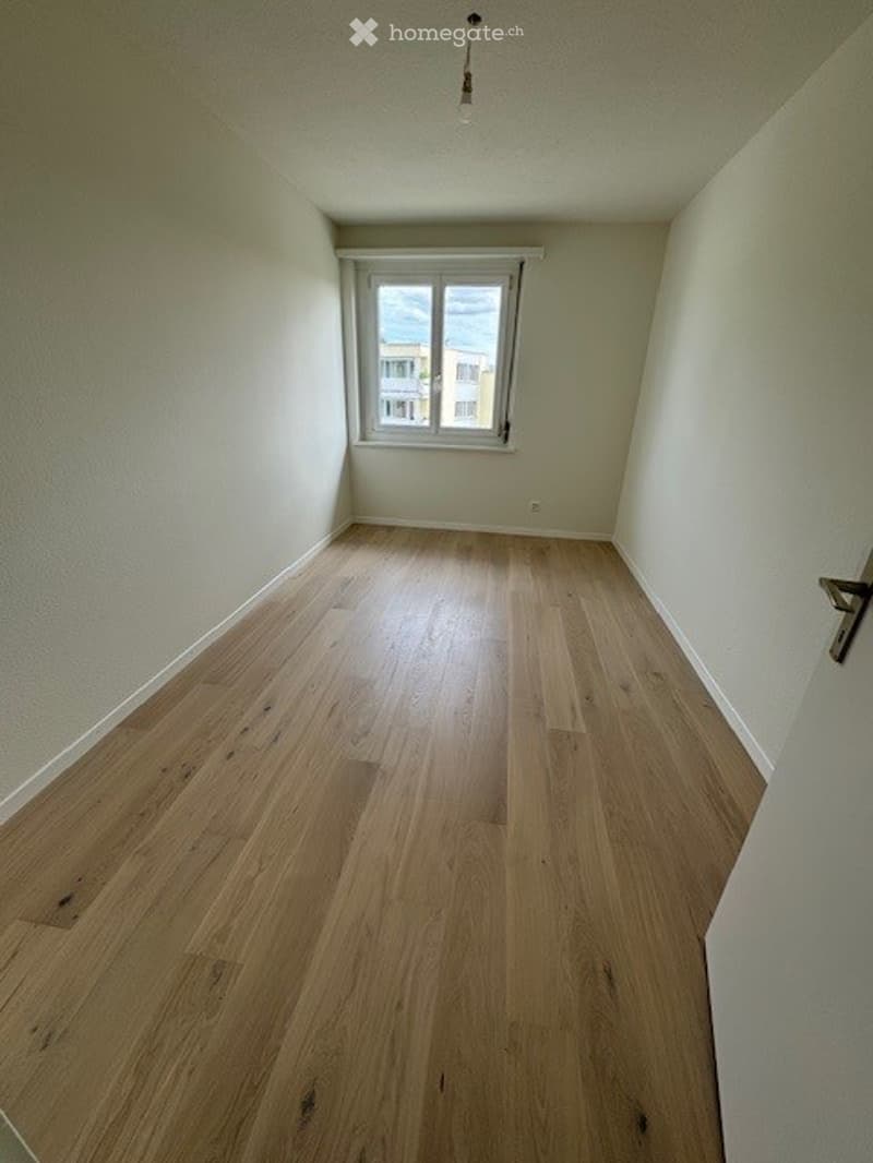 Empty room with light wooden flooring and white walls, featuring a single window with white frame and a door visible on the right side.