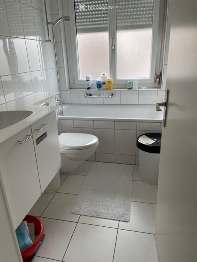 Modern bathroom with white tiles, a bathtub, toilet, sink, and a small rug on the floor. A window with frosted glass is above the bathtub, and a few toiletries are placed on the windowsill.