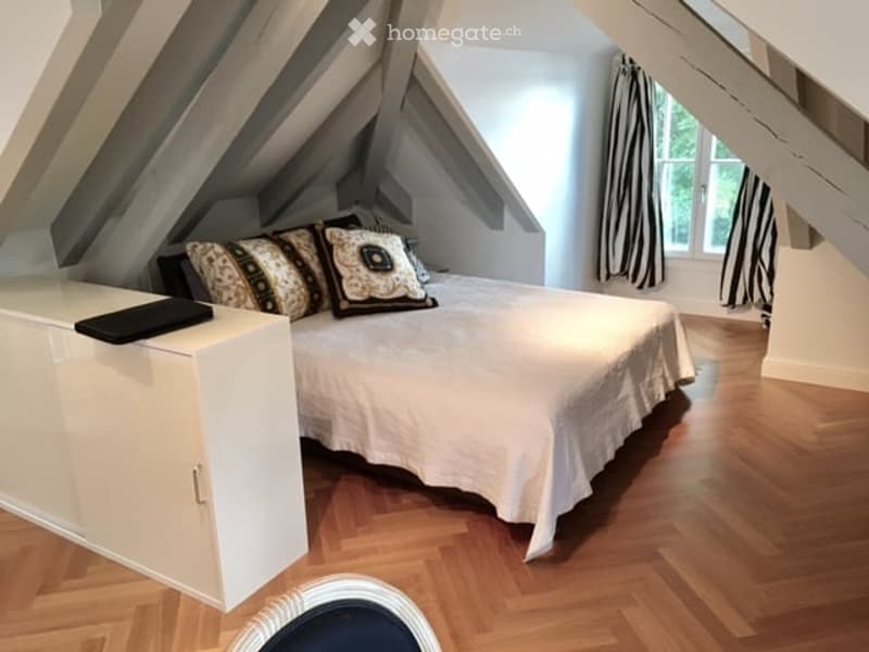 Cozy attic bedroom with wooden flooring, slanted ceilings, large window with striped curtains, neatly made bed with decorative pillows, and a white cabinet with a laptop on top.