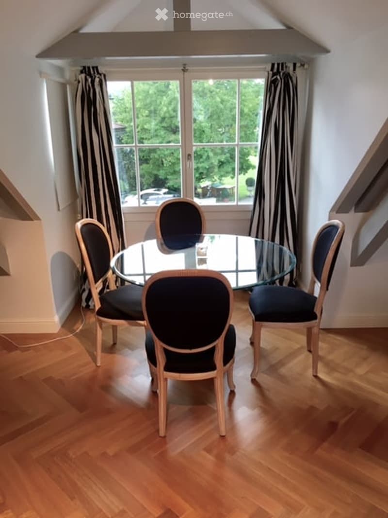 A small, well-lit dining area with a round glass table and four matching chairs, set against a backdrop of large windows with striped curtains. The room features wooden flooring and a sloped ceiling typical of an attic space.