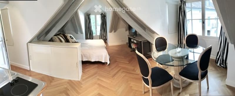 Cozy attic room with wooden herringbone flooring, a bed, a small dresser, a glass dining table with chairs, and a television on a stand.