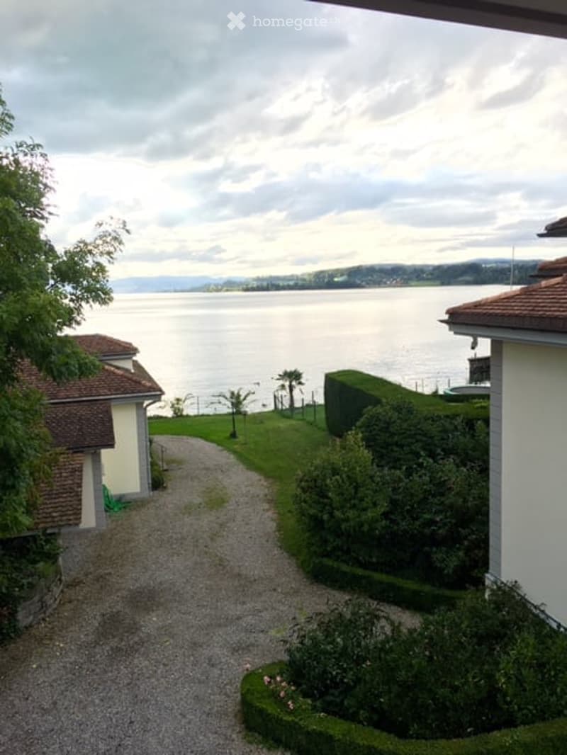 Garden area with a view of a lake, gravel pathway, trimmed hedges, and surrounding greenery.