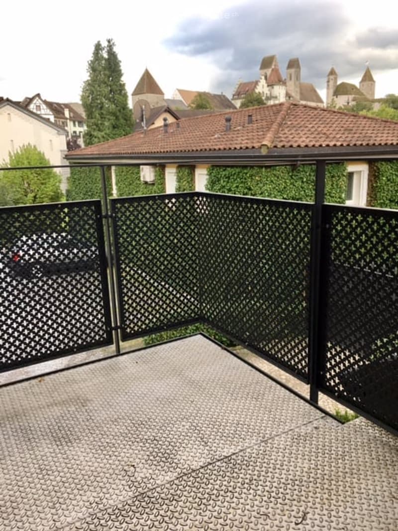 Balcony with black metal lattice fencing, grey tiled floor, overlooking a historic town with visible rooftops and a castle in the background.