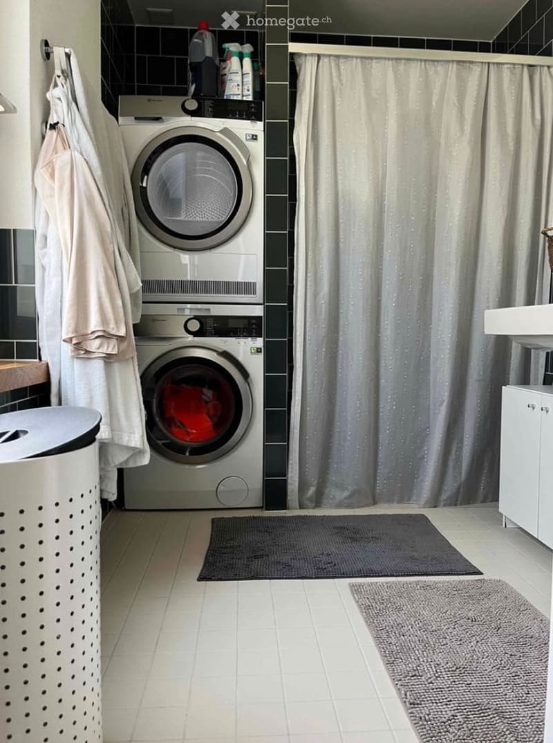Modern bathroom with a stacked washer and dryer unit, a shower curtain, towels, cleaning supplies, a trash bin, and a bathroom cabinet.