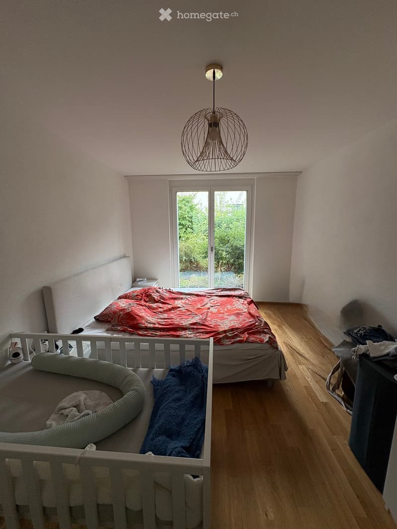 Cozy bedroom with a double bed, a cat bed, a nightstand, and a modern hanging lamp. The room features wooden flooring and large windows with a view of greenery.