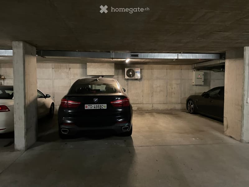 Underground parking area with three parked cars, concrete pillars, and an air conditioning unit mounted on the wall.