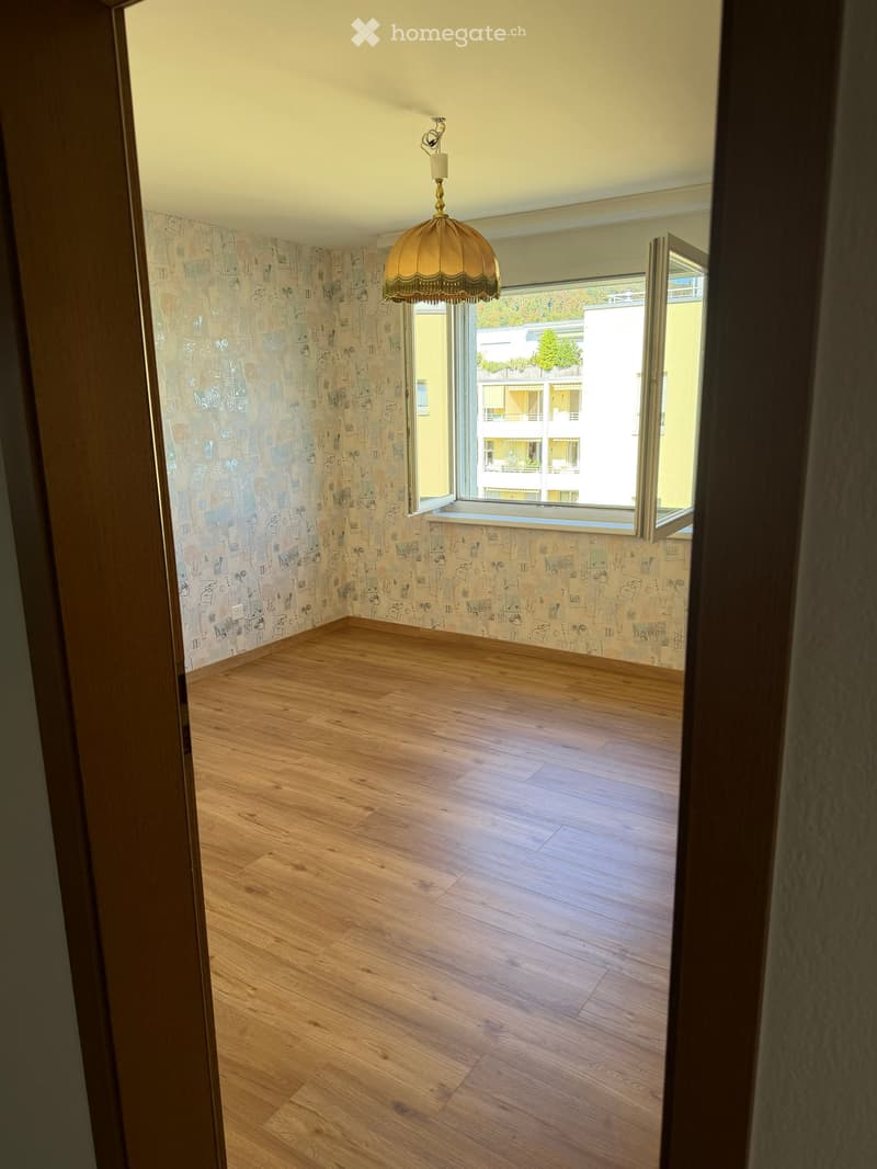 Empty room with wooden flooring, patterned wallpaper, and a large window with a view of neighboring buildings.