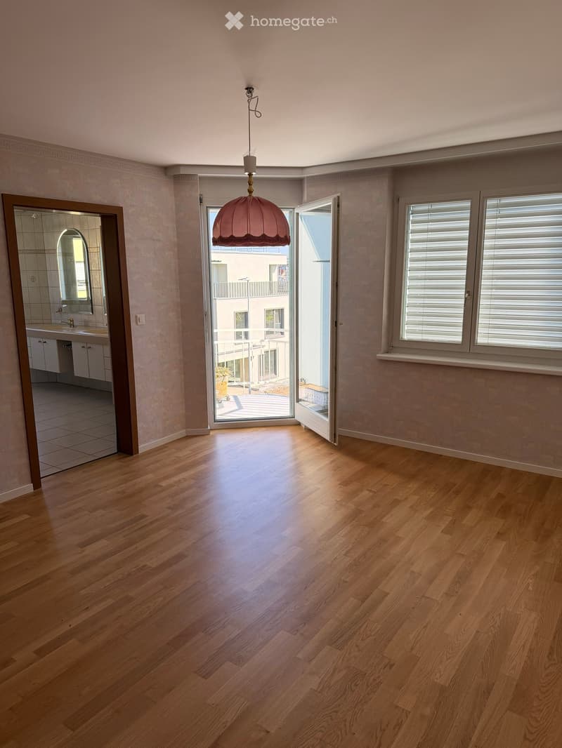 Empty room with wooden flooring, large windows with blinds, and an open door leading to a balcony. A chandelier hangs from the ceiling, and a small bathroom is visible to the left.