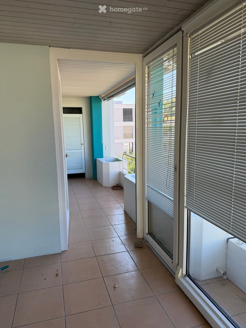 Balcony with tiled floor, white walls, large windows with blinds, and a small white planter on the left side.
