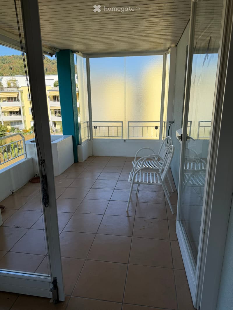 Balcony with tiled floor, white metal chairs, glass railings, and a view of surrounding buildings and greenery.