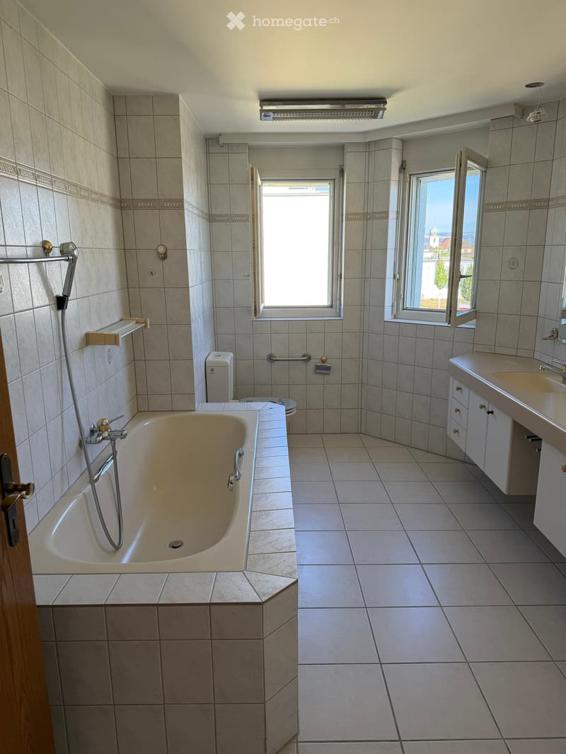 Modern bathroom with tiled walls and floor, featuring a bathtub, sink, toilet, and large windows offering a view.