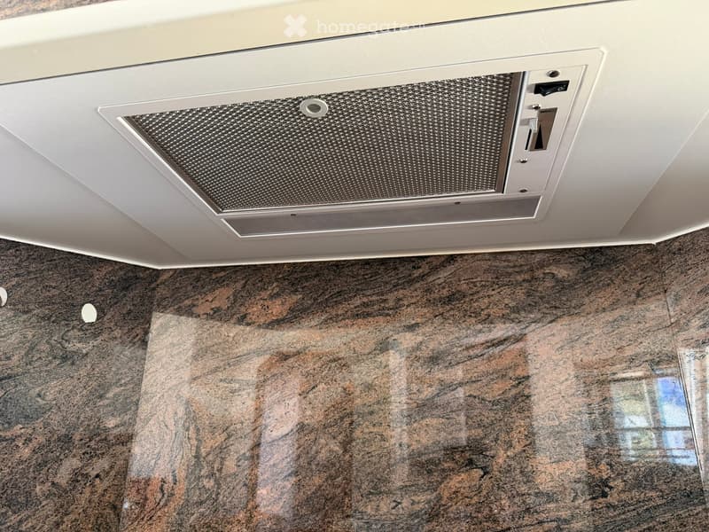 Close-up view of a kitchen ceiling showing a white range hood with a mesh filter and control panel, above a dark granite countertop.