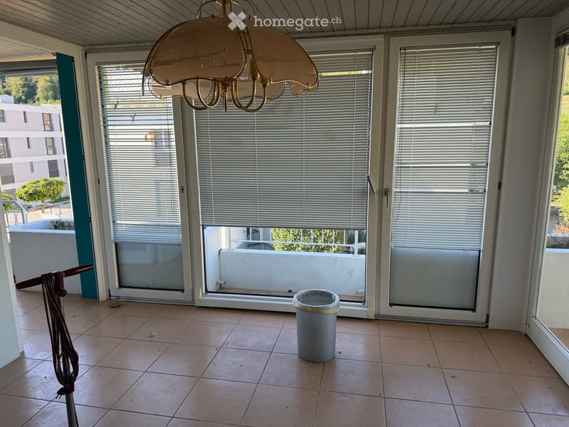 Empty room with tiled floor, large sliding glass doors with blinds, chandelier, broom, and trash bin.