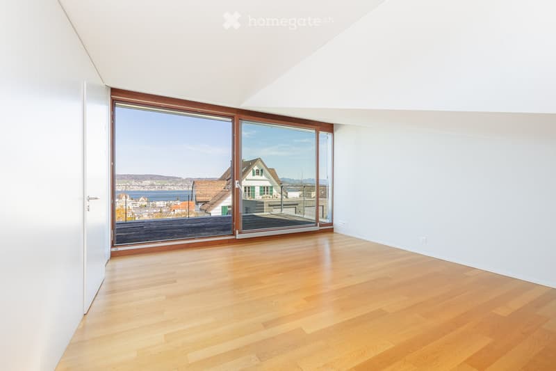 Empty room with wooden floors, white walls, and large windows with a view of the city and lake.