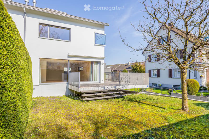 Modern white house with glass windows and a wooden deck, surrounded by a well-maintained garden.