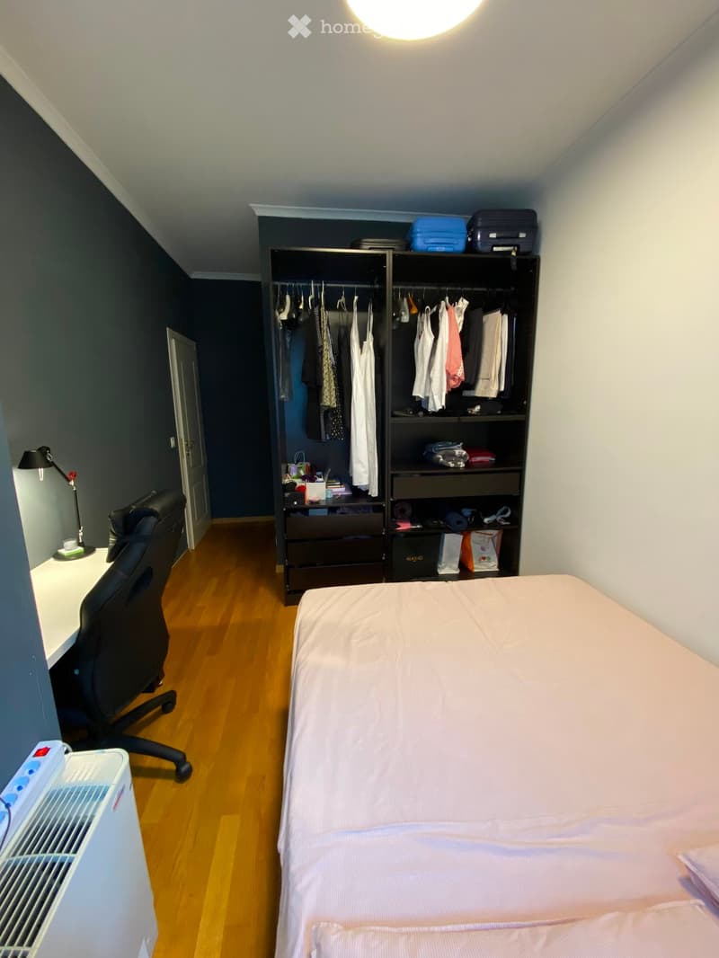 A modern bedroom with a neatly made bed, a wardrobe with hanging clothes, a desk with a chair, and a radiator on the left side.