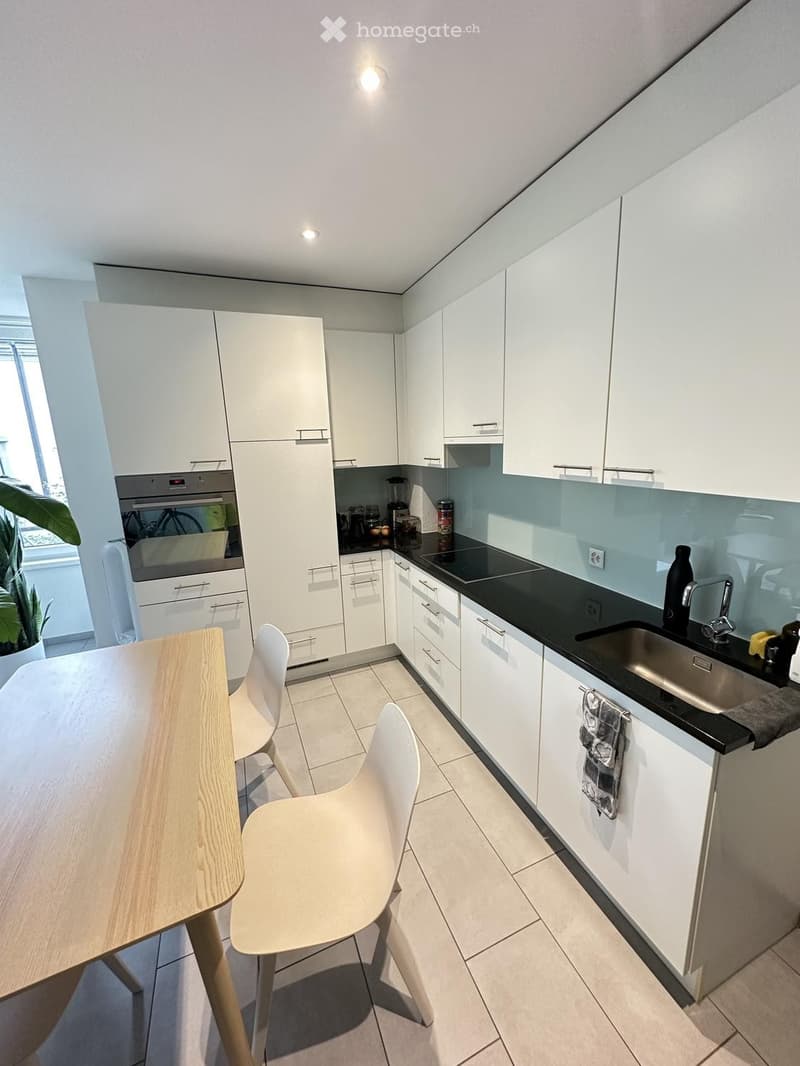 Modern kitchen with white cabinets, black countertops, a wooden dining table with white chairs, and various appliances including an oven, sink, and blender.
