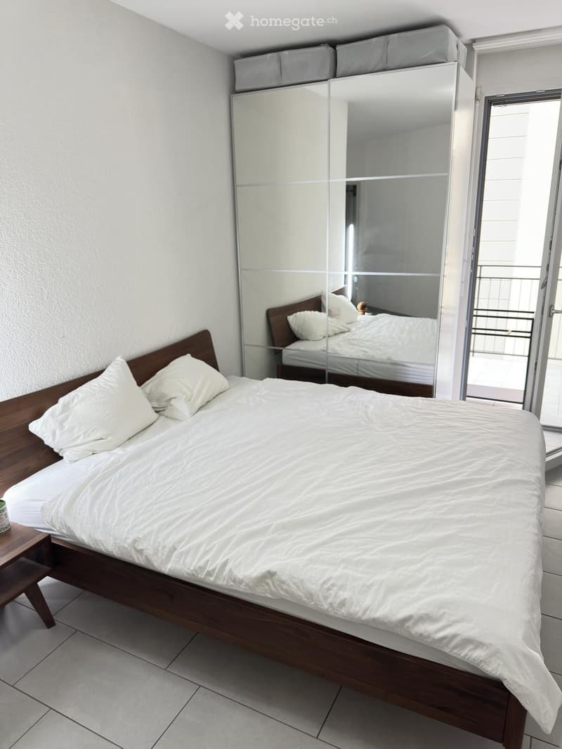 A modern bedroom with a wooden bed, white bedding, a large mirrored wardrobe, and a small nightstand.