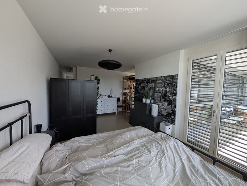 Modern bedroom with a large bed, grey bedding, a black wardrobe, a nightstand, a chair, a shelf, and a table. The room features large windows with blinds and a view of the city.