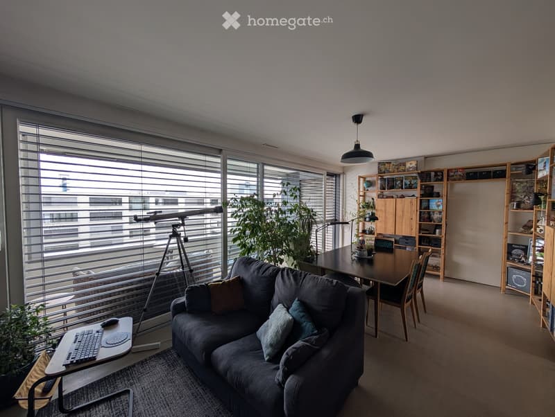 Modern living room with large windows, grey sofa, laptop on side table, dining table with chairs, bookshelf, and potted plants.
