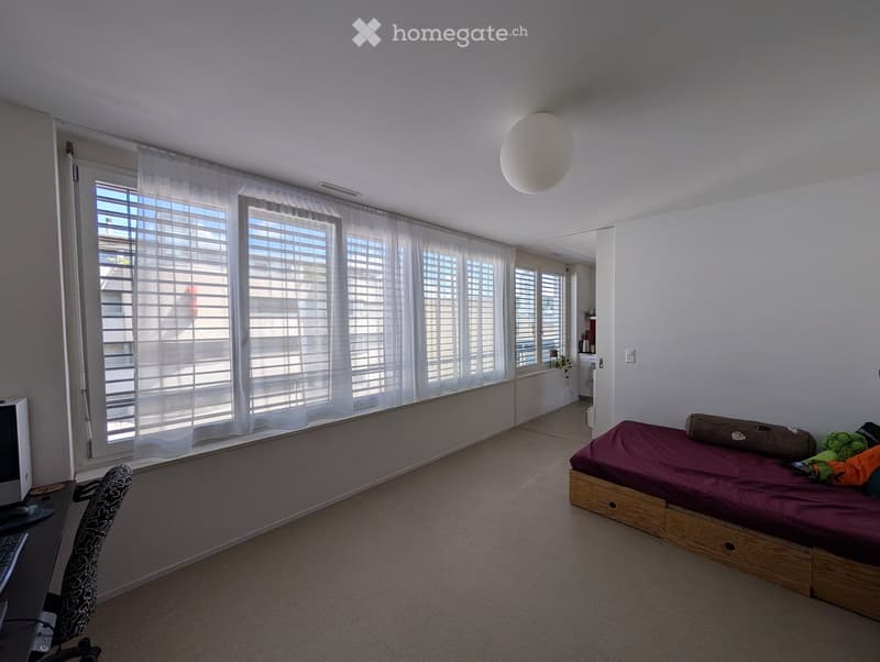 A furnished bedroom with a bed, a chair, a computer setup, and large windows with white curtains.