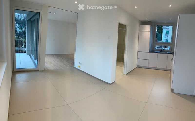 Empty room, sliding glass doors leading to balcony, tiled floor, white walls, recessed ceiling lights, open kitchen with white cabinets, refrigerator, countertop with sink