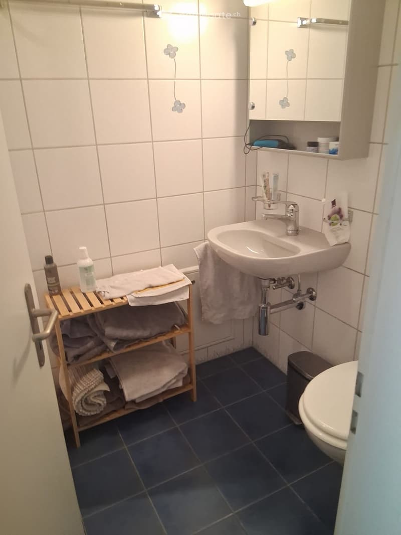 A small bathroom with white tiled walls, a white sink with a silver faucet, a mirror above the sink, a wooden towel rack with folded towels, a toilet, and a small cabinet with toiletries.