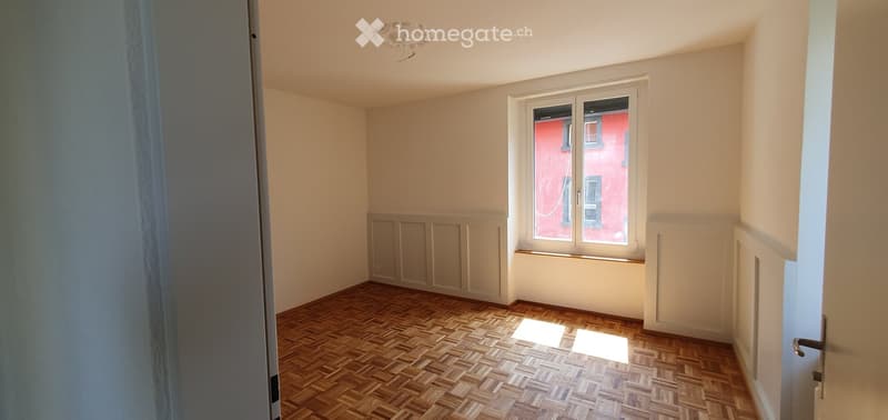 Empty room with wooden parquet flooring, white walls, and a large window with a view of a red building. The room features a simple, clean design with no furniture or decorations.