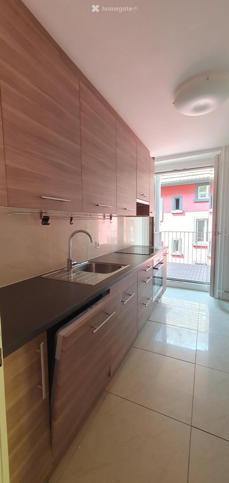 Modern kitchen with wooden cabinets, stainless steel sink, and countertop, leading to a balcony area.