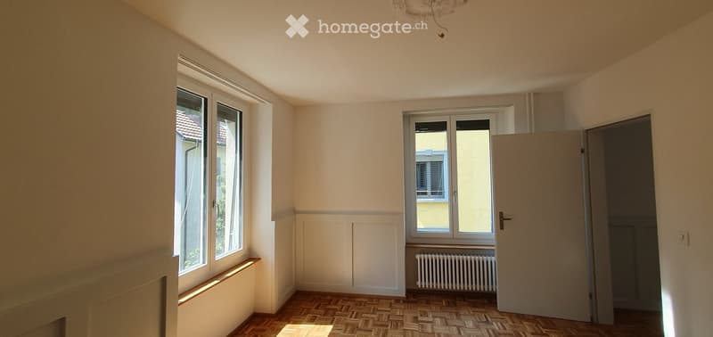 Empty room with wooden flooring, white walls, large windows, and a radiator. No furniture or equipment present.