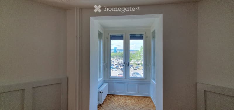 Empty room with wooden herringbone flooring, white walls, large windows showing a street view, and a radiator on the left side.