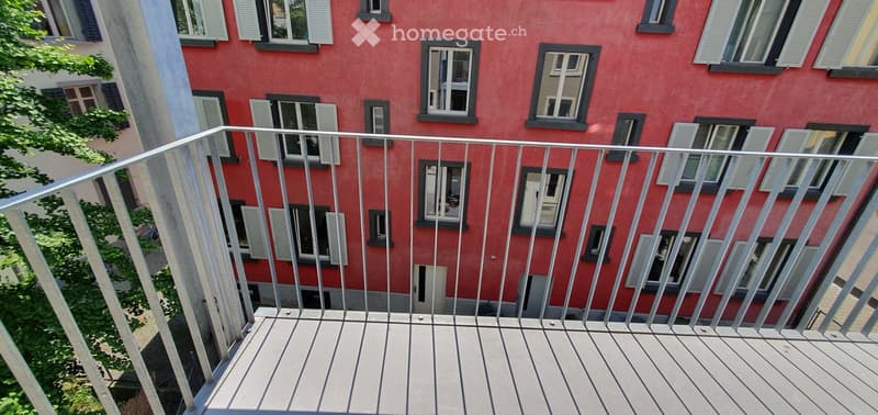 Balcony with wooden decking and metal railing, overlooking a red brick building with multiple windows and shutters.
