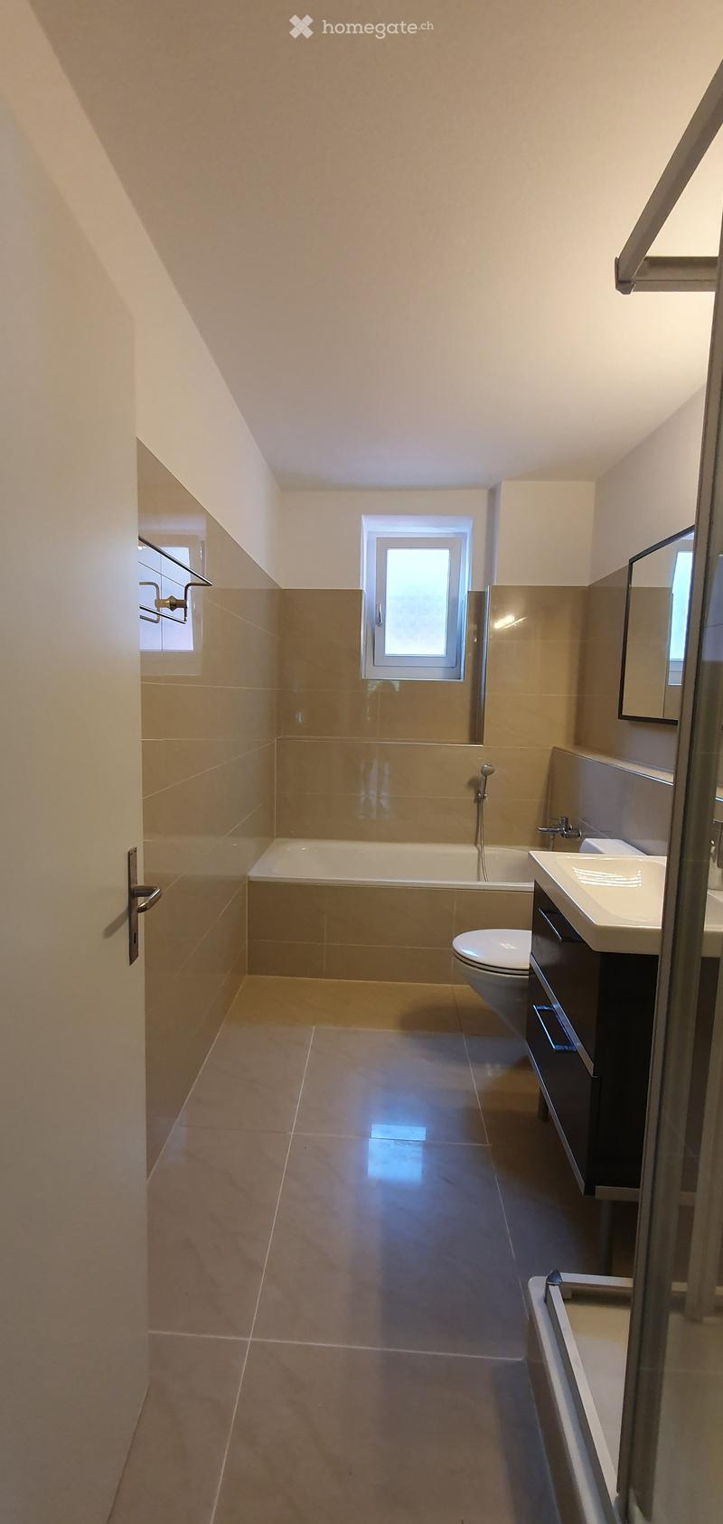 Modern bathroom with beige tiles, a bathtub, toilet, sink, and a large mirror. Features a window and a glass shower enclosure.
