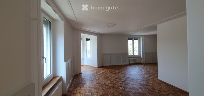 Empty room with wooden parquet flooring, white walls, and large windows allowing natural light. The room features a simple ceiling light fixture and a radiator under one of the windows.