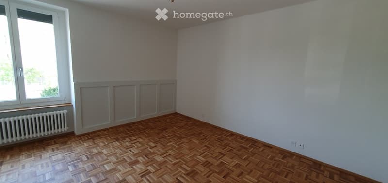 Empty room with wooden parquet flooring, white walls, and a large window with a radiator below it.