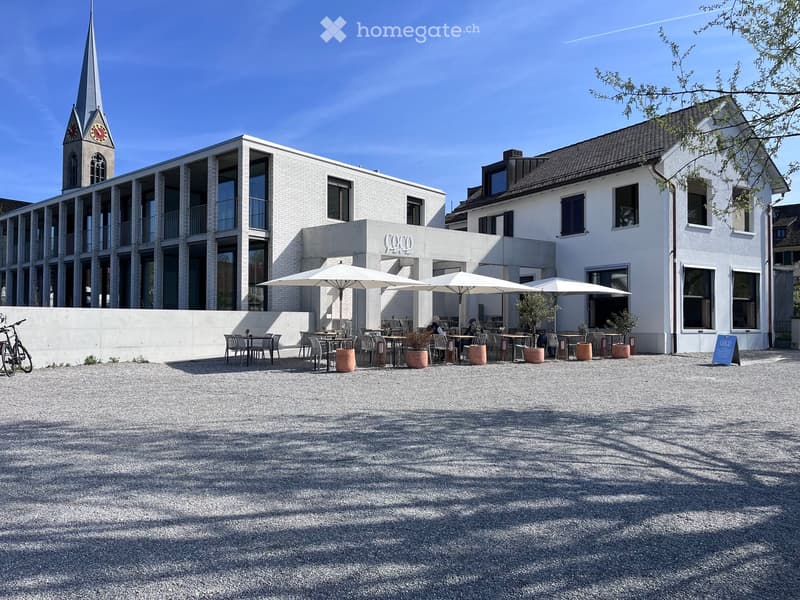 Courtyard area with outdoor seating, umbrellas, tables, and chairs, surrounded by modern buildings and a church tower.