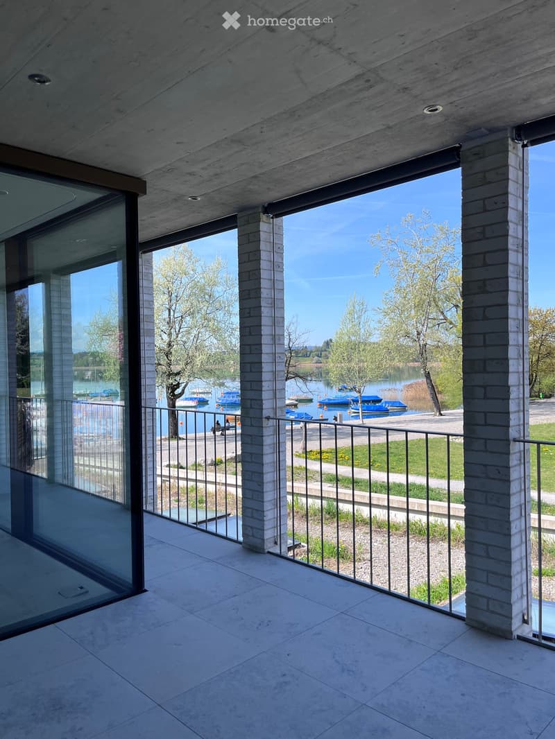 Modern balcony with large glass panels, white brick columns, and a metal railing, overlooking a lake with boats and trees.